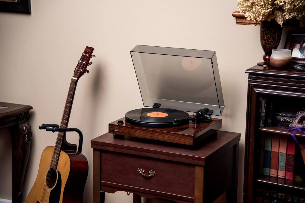 Wood-finished turntable with a transparent dust cover, playing a vinyl record on a wooden side table, next to an acoustic guitar and a bookshelf in a cozy living room.