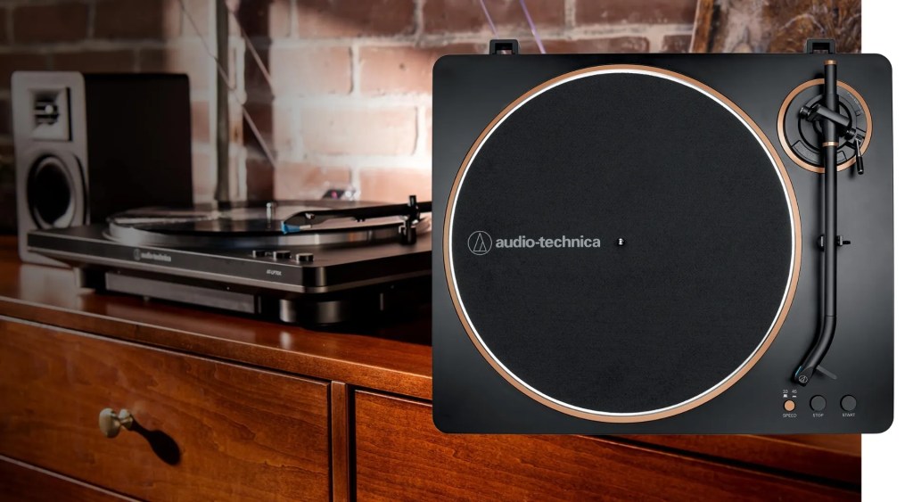 Audio-Technica LP-70X turntable on a wooden dresser and a rustic brick wall in the background. The turntable features a matte black finish with copper accents and is spinning a vinyl record.