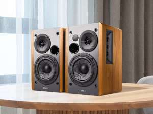 Two Edifier R1280T bookshelf speakers with a classic wood veneer finish and black front panels are displayed on a light wood table. The right speaker features volume, bass, and treble control knobs on the side. Sheer curtains and gray drapes are visible in the softly lit background.