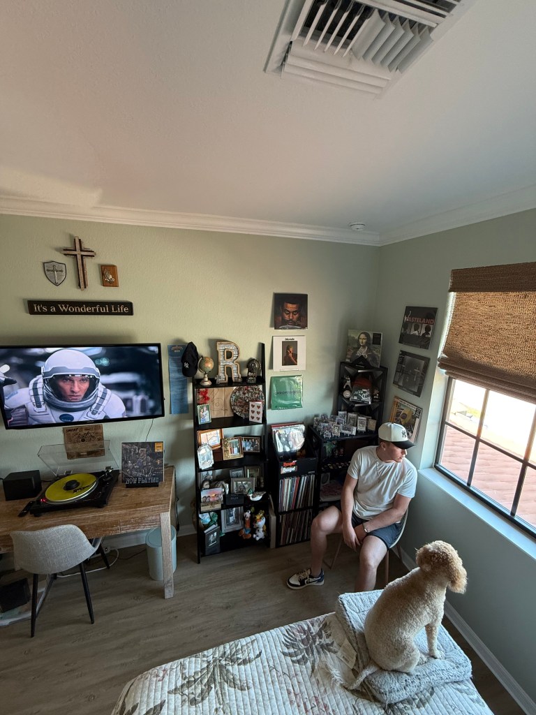 The same man sits in the corner of his listening room, facing a shelf stacked with records, personal memorabilia, and a small TV screen showing a scene from “Interstellar.” A dog lies peacefully on the bed in the foreground, adding a relaxed, homey vibe.