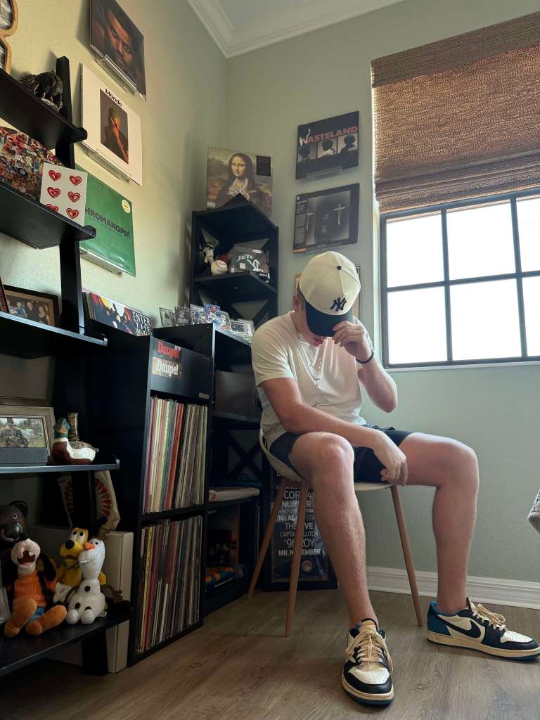 A man in a white t-shirt, navy shorts, and a Yankees cap sits on a wooden chair in a vinyl-themed room. Behind him are shelves filled with records, collectibles, and framed art. Plush toys and vinyl accessories are visible, adding character to the cozy space.