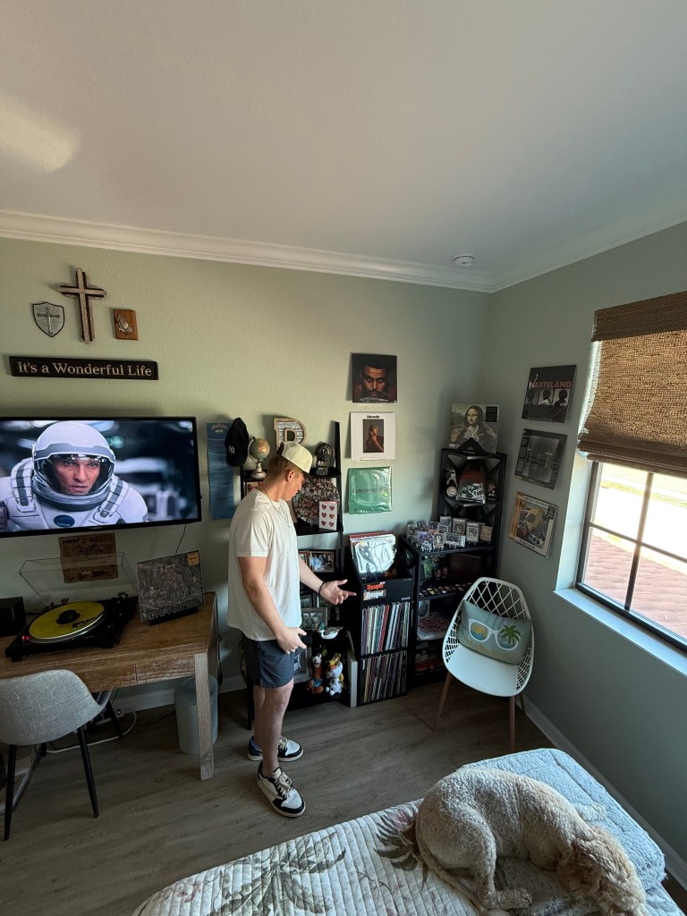 The man stands and gestures toward his shelf, filled with vinyl records, framed photos, and unique decor. A turntable setup is visible on a nearby desk, and a white accent chair is tucked near the window. Natural light streams into the inviting, music-filled room.