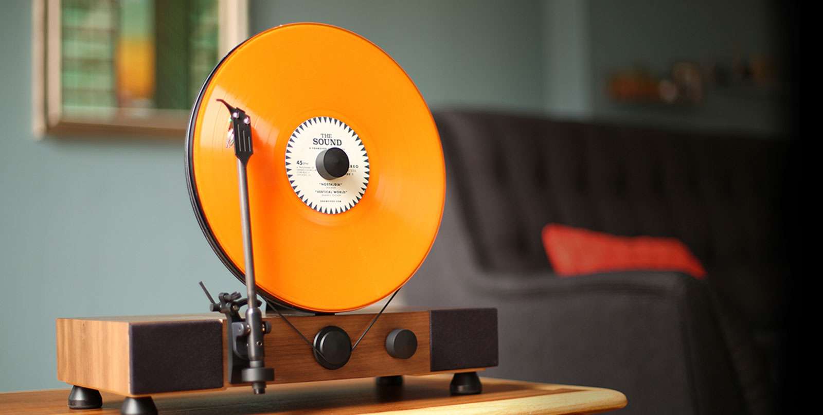 A close-up side angle view of a vertical record player with an eye-catching orange vinyl record, mounted upright on a minimalist wooden base, featuring black speakers and control knobs, set in a stylish and cozy interior.