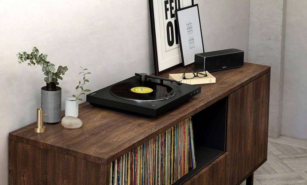 A black Sony PS-LX310BT belt-drive turntable playing a vinyl record on a dark wood console; to the left sits a matching black Sony wireless speaker, with a potted plant and framed vinyl cover resting behind.