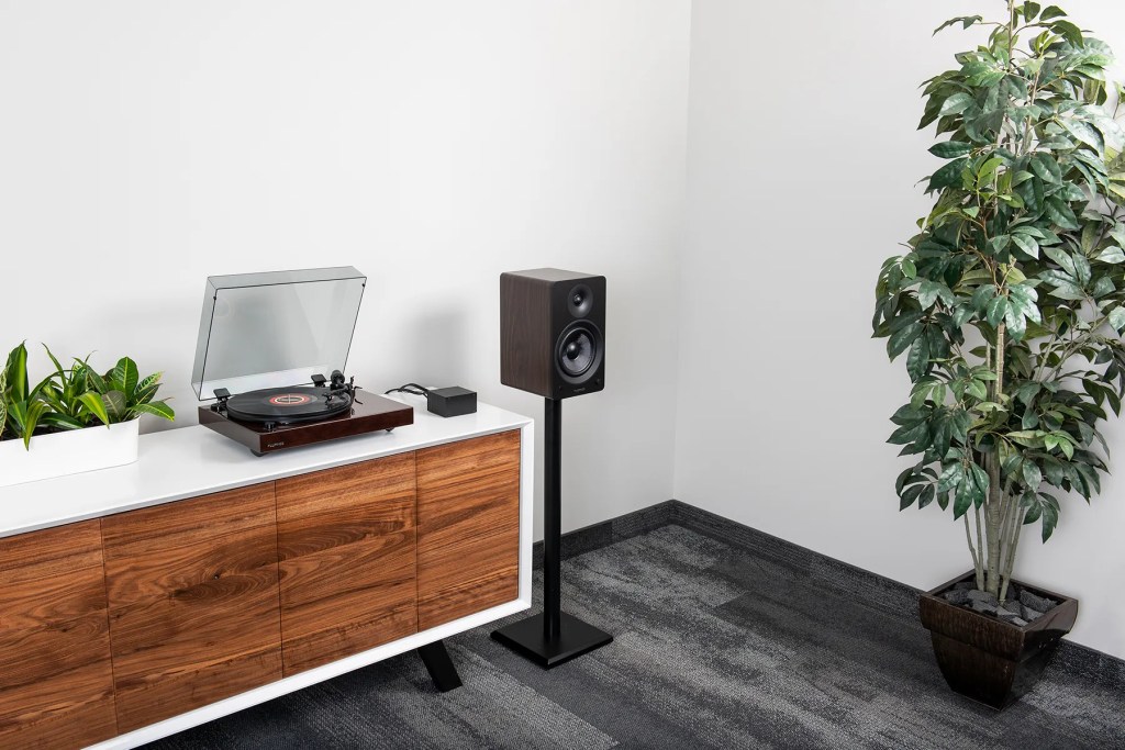 A minimalist white-and-walnut media console topped by a walnut-finish turntable with its clear dust cover open, a small black power supply box, and a white planter of green houseplants; to the right, a matching walnut-finish bookshelf speaker sits on a tall black stand against a white wall and gray carpet, with a potted ficus in a dark wooden planter in the corner.