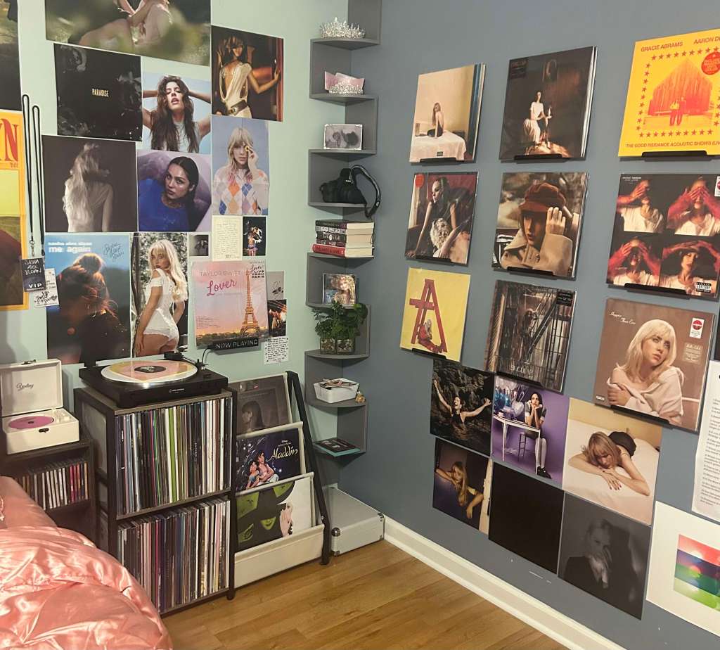 A cozy bedroom vinyl setup featuring two stacked record shelves filled with LPs, a Crosley turntable with a pink vinyl spinning, and a white mini CD player. The walls are decorated with posters and framed album covers of artists like Taylor Swift, Billie Eilish, and others, creating a personalized music corner with a mix of pop culture and personal touches.