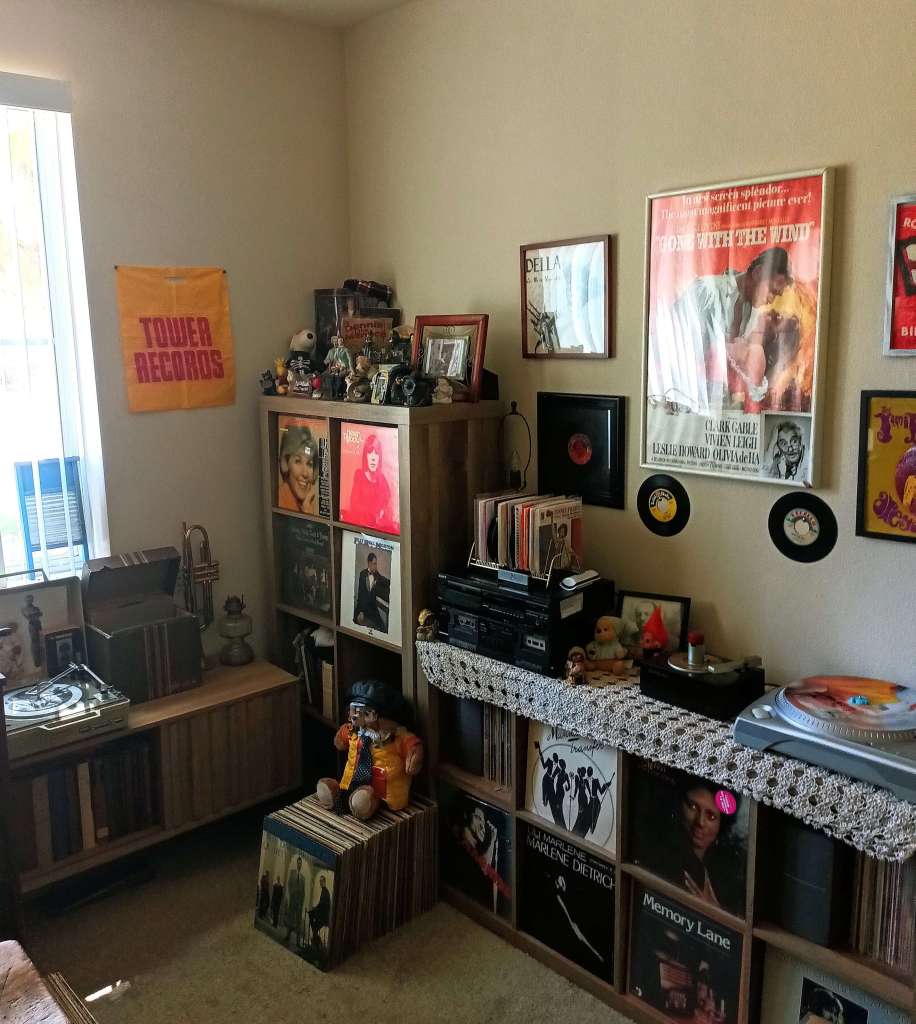 Cozy vintage vinyl listening room filled with wooden record shelves, a turntable on the left, stereo gear on a lace-covered cabinet, framed posters and album art on the walls, and a nostalgic Tower Records bag hanging near the window.