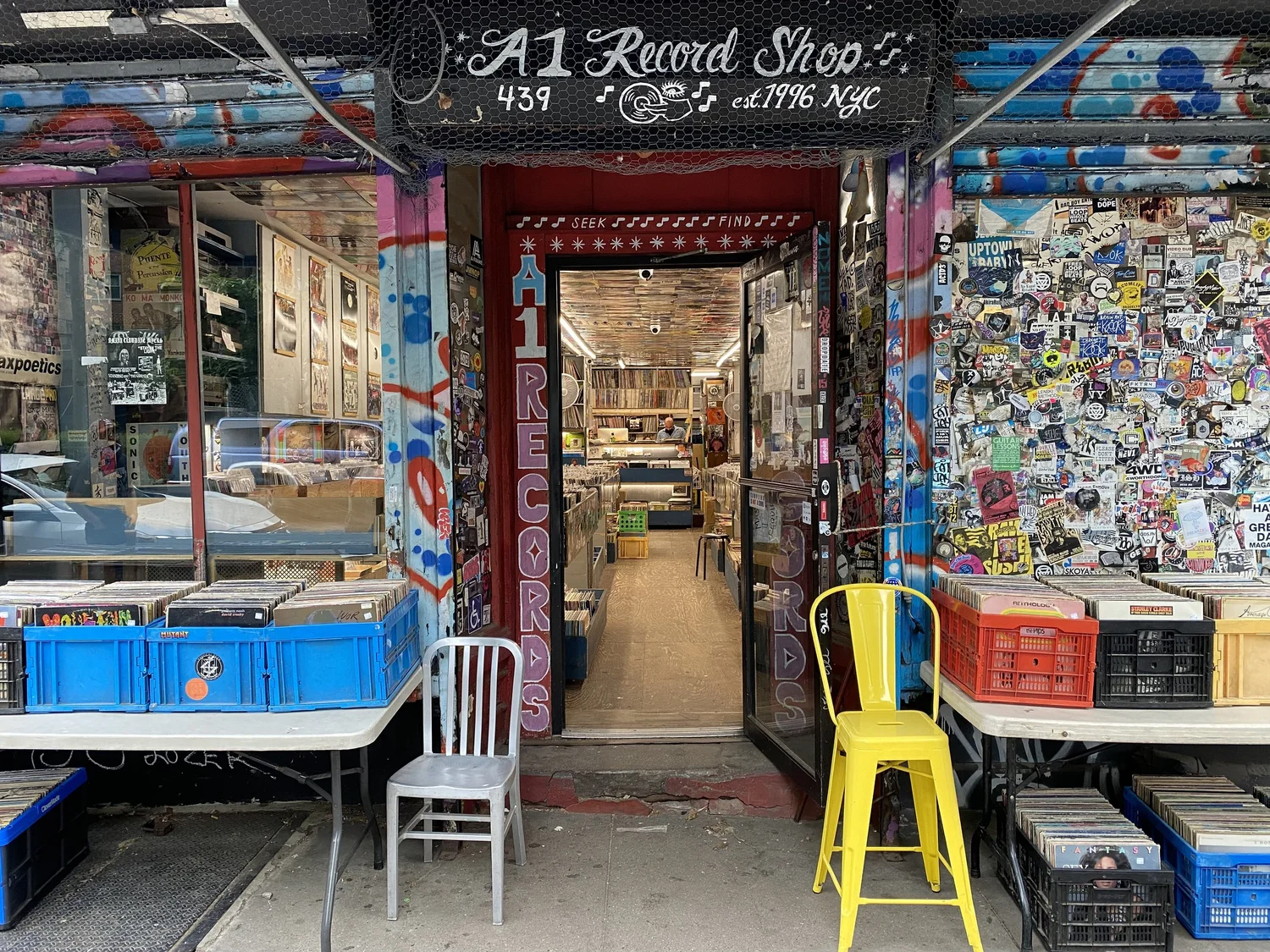 Exterior of A1 Record Shop in New York City, with a colorful graffiti-covered storefront, sticker-covered wall, and open doorway leading into rows of vinyl inside. Crates of records are set out on tables and in bins on the sidewalk, with metal chairs—including a bright yellow one—placed out front.