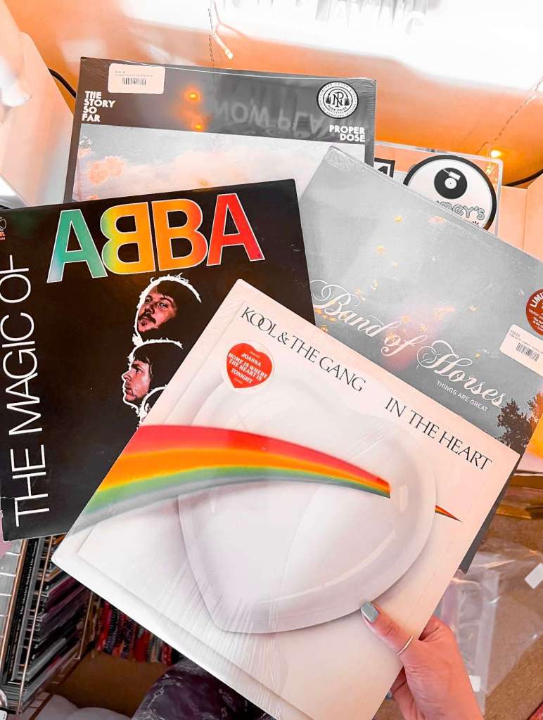 Hand holding a stack of vinyl records including ABBA, Kool & The Gang, Band of Horses, and The Story So Far, with warm lighting in the background.