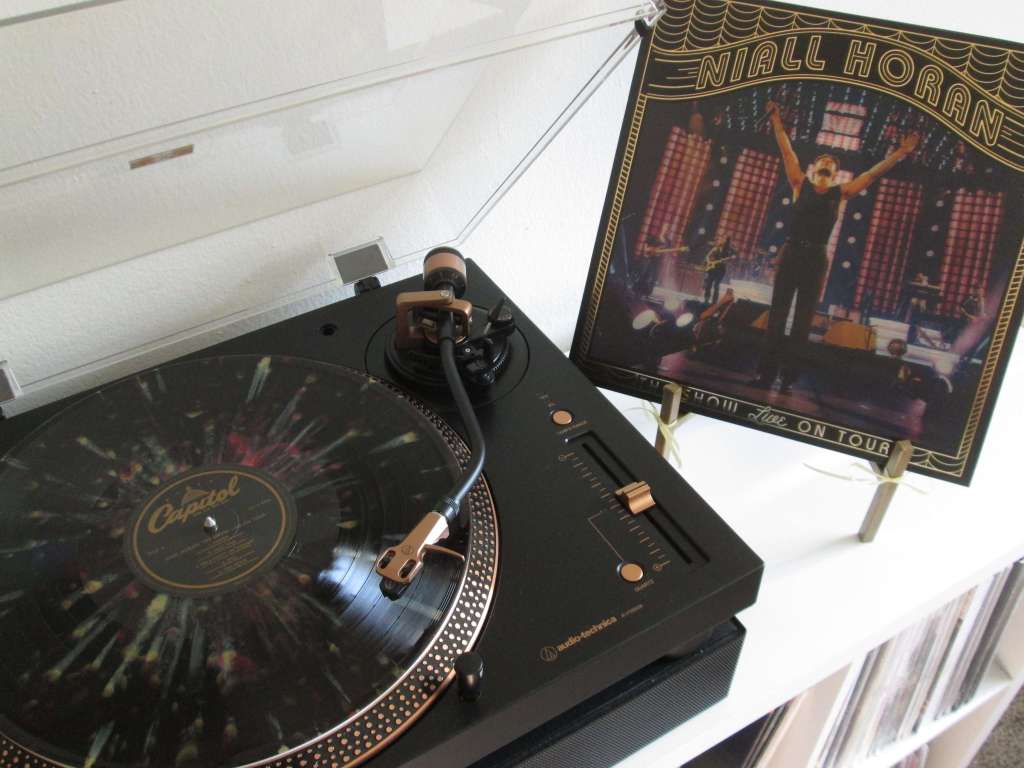 A Niall Horan “The Show Live on Tour” vinyl record spinning on an Audio-Technica turntable with bronze accents. The record has a colorful splatter design, and the album cover is propped up on a gold easel next to the turntable.