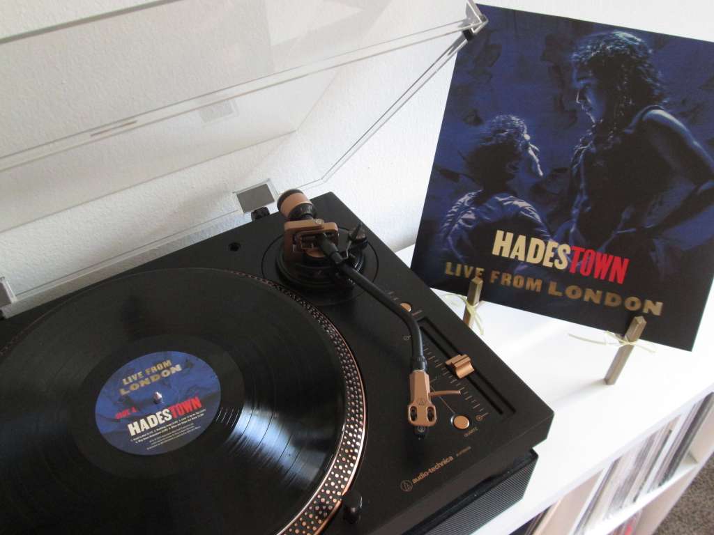 A Hadestown “Live from London” vinyl record playing on an Audio-Technica turntable with bronze accents. The record is black with a custom center label, and the album cover featuring two characters facing each other is displayed on a gold easel beside the turntable.