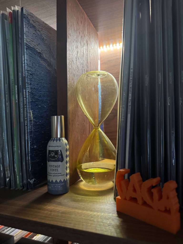 Close-up of a vinyl shelf display featuring a yellow glass hourglass, a Vintage Vinyl room spray, and an orange 3D-printed GAGA sign, with records stacked on either side.