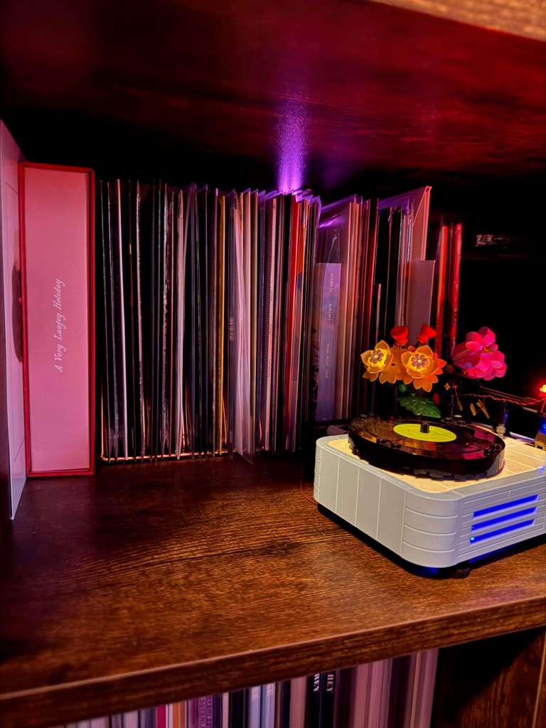 A cozy wooden shelf filled with vinyl records and a small Lego record player topped with a black disc and a bouquet of Lego flowers. Warm pink and purple lighting casts a soft glow across the spines of the records.
