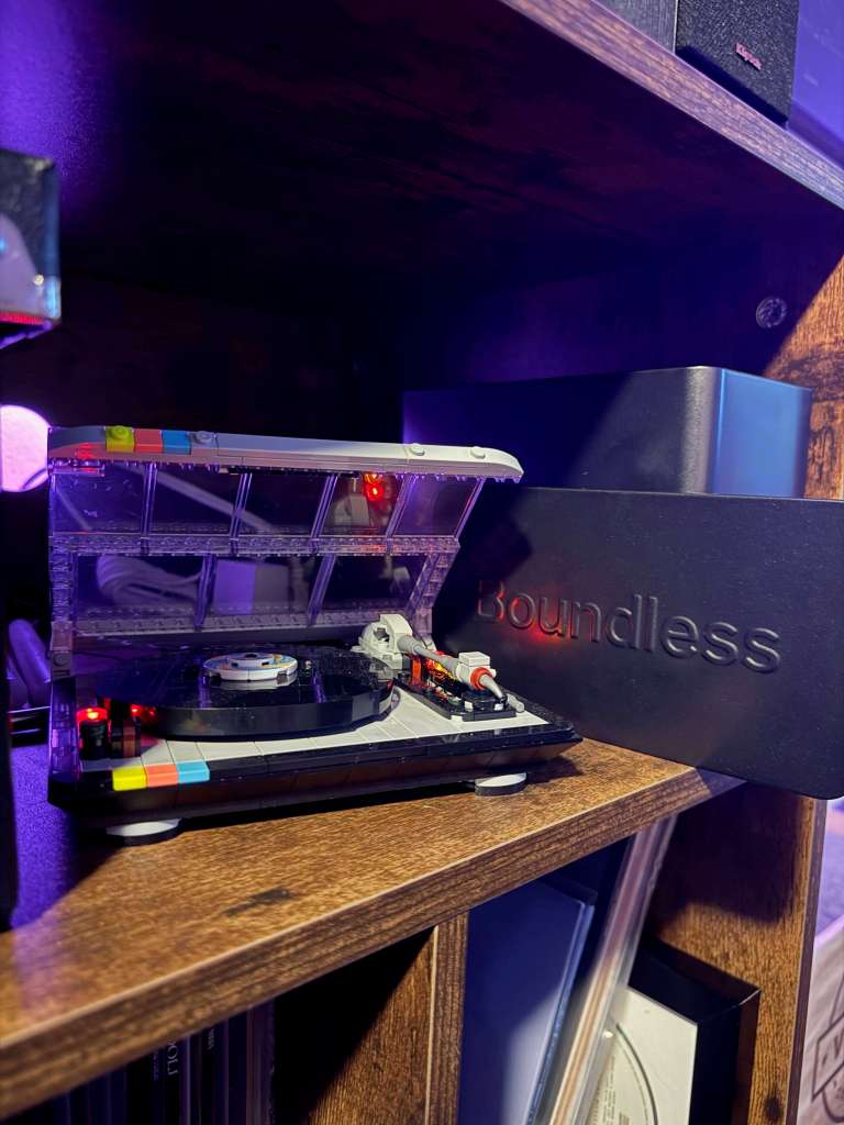 A close-up of a miniature Lego turntable with a clear lid, colorful buttons, and a spinning record, sitting on a wooden shelf beside a black Boundless box. Soft purple lighting gives the setup a futuristic glow.