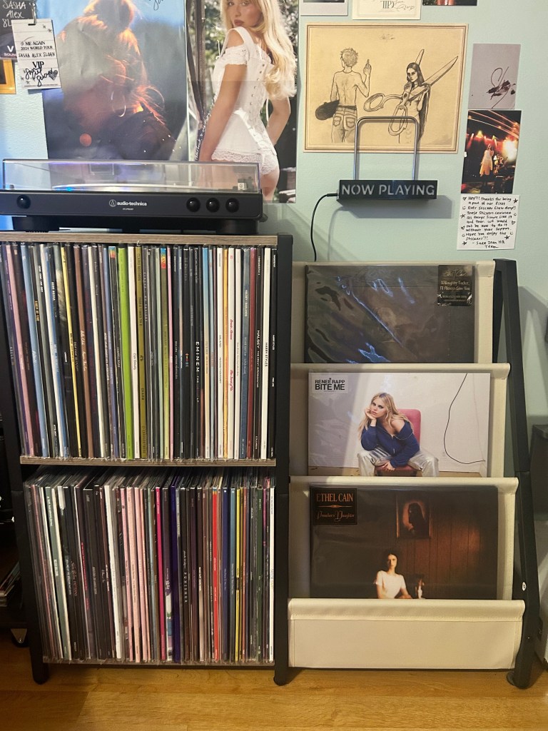 A close-up of Cass’s record storage with two shelves packed full of vinyl albums. Next to it, a three-slot stand displays feature records, including Renee Rapp – Bite Me and Ethel Cain – Preacher’s Daughter. A “Now Playing” sign is mounted on the wall above.
