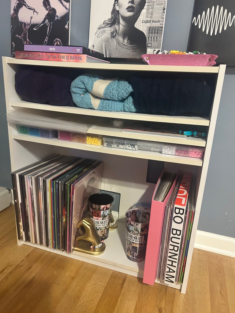 A white shelving unit holding vinyl records on the bottom shelves, alongside Taylor Swift tour cups and a gold hand bookend. The middle shelves store folded blankets, while the top has colorful craft beads and books, including Olivia Rodrigo’s Guts.
