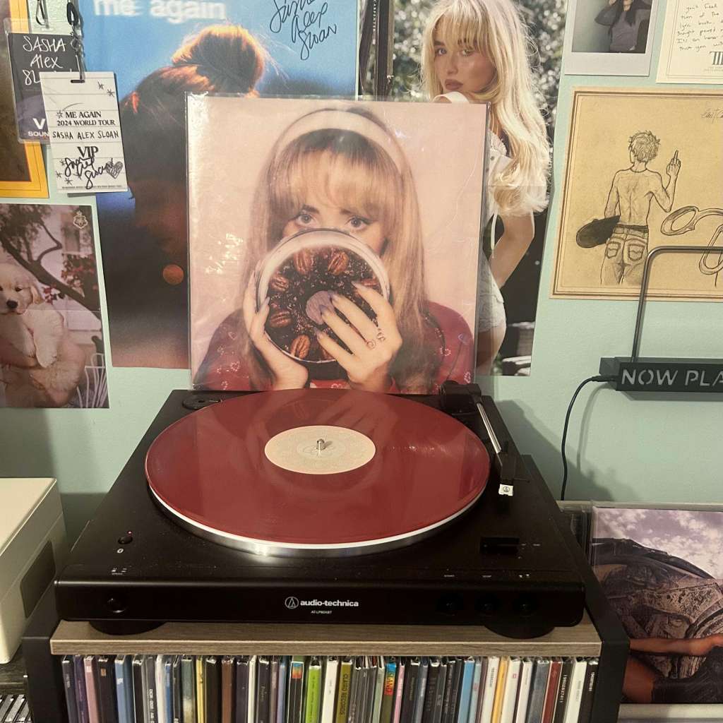 A deep red vinyl record spinning on an Audio-Technica LP60X, with album art showing a woman holding a pie in front of her face in the background.