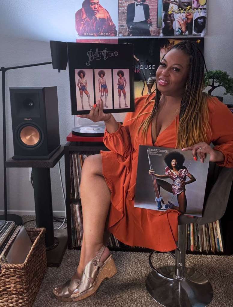 Alt text: A woman in an orange dress sits in her listening room holding two Betty Davis records, one in each hand. Behind her are wall-mounted vinyl covers including D’Angelo’s Brown Sugar and Amy Winehouse’s Back to Black. A Klipsch speaker, red turntable, and rows of vinyl records are visible around her setup.