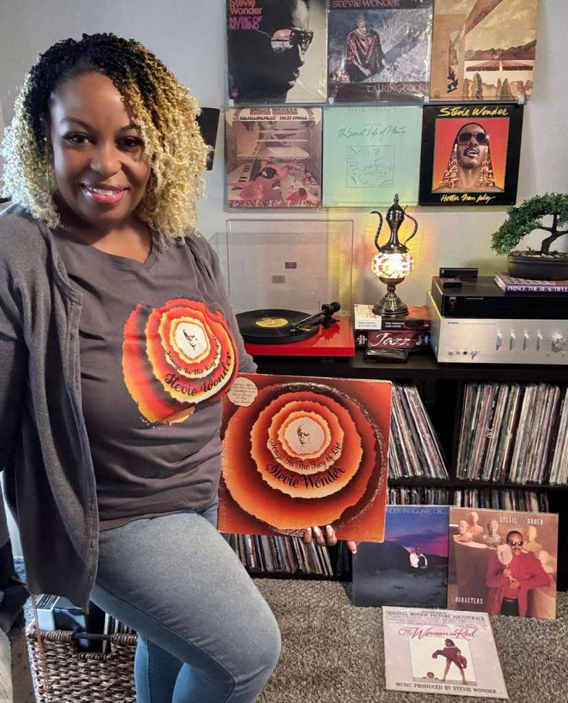A woman wearing a Stevie Wonder t-shirt poses in front of her turntable setup, proudly holding the album Songs in the Key of Life. Behind her, multiple Stevie Wonder records are displayed on the wall. Her setup includes a red turntable, Yamaha amplifier, bonsai plant, and mosaic lamp with shelves full of vinyl below.