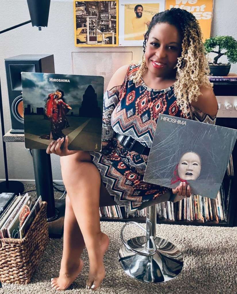 A woman in a patterned dress sits barefoot in her listening room, holding two Hiroshima records in her hands. Behind her are wall-mounted vinyl covers including Roy Ayers and Donald Byrd. Her setup features a red turntable, Klipsch speakers, a bonsai plant, and shelves overflowing with records.
