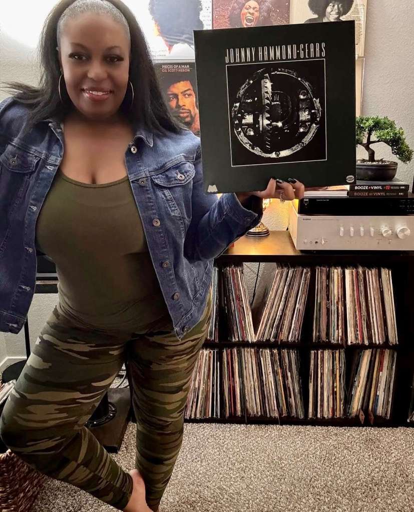 A woman in a denim jacket and camouflage pants stands next to her vinyl setup, holding Johnny Hammond’s Gears album. Behind her are records on display including Gil Scott-Heron’s Pieces of a Man. Her shelves are stacked with vinyl, alongside a modern hi-fi setup.