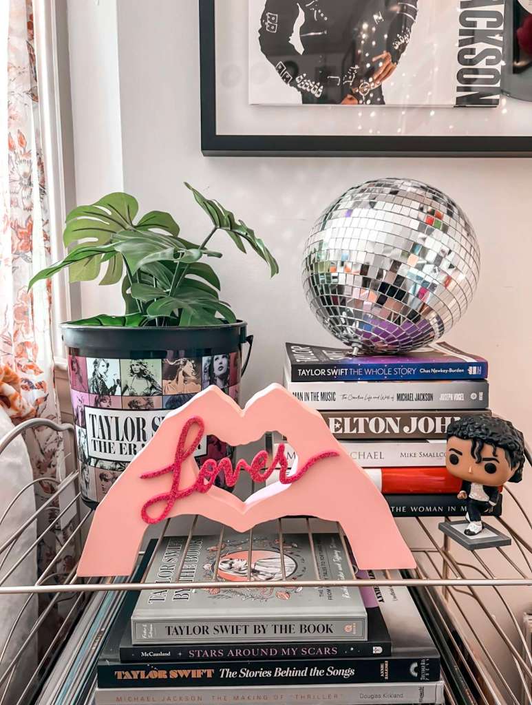 A stylish shelf stacked with music books, including several Taylor Swift titles, Elton John, and Michael Jackson. A silver disco ball, leafy plant, and a pink “Lover” sign shaped like lips sit on top. Funko Pop figurines add a playful touch, tying into the music-themed decor.