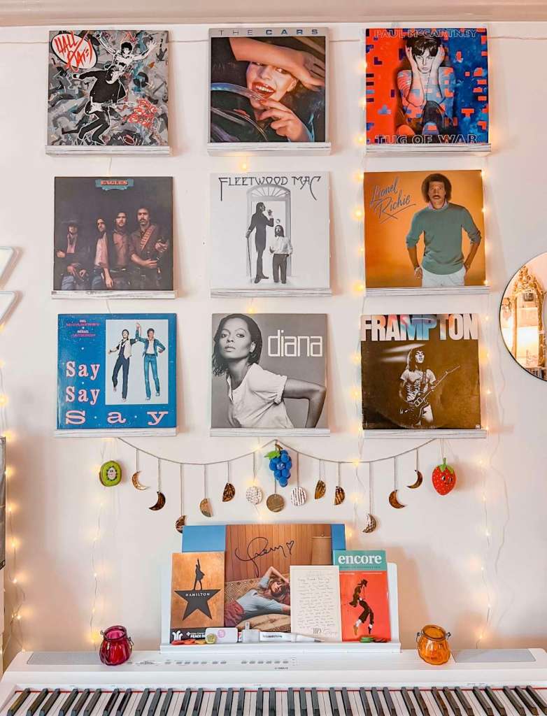 A wall display of framed vinyl albums, including Hall & Oates, The Cars, Paul McCartney, Eagles, Fleetwood Mac, Diana Ross, and Peter Frampton. Below is a white keyboard decorated with candles, postcards, and a small signed note, with string lights and hanging celestial garlands adding warmth.