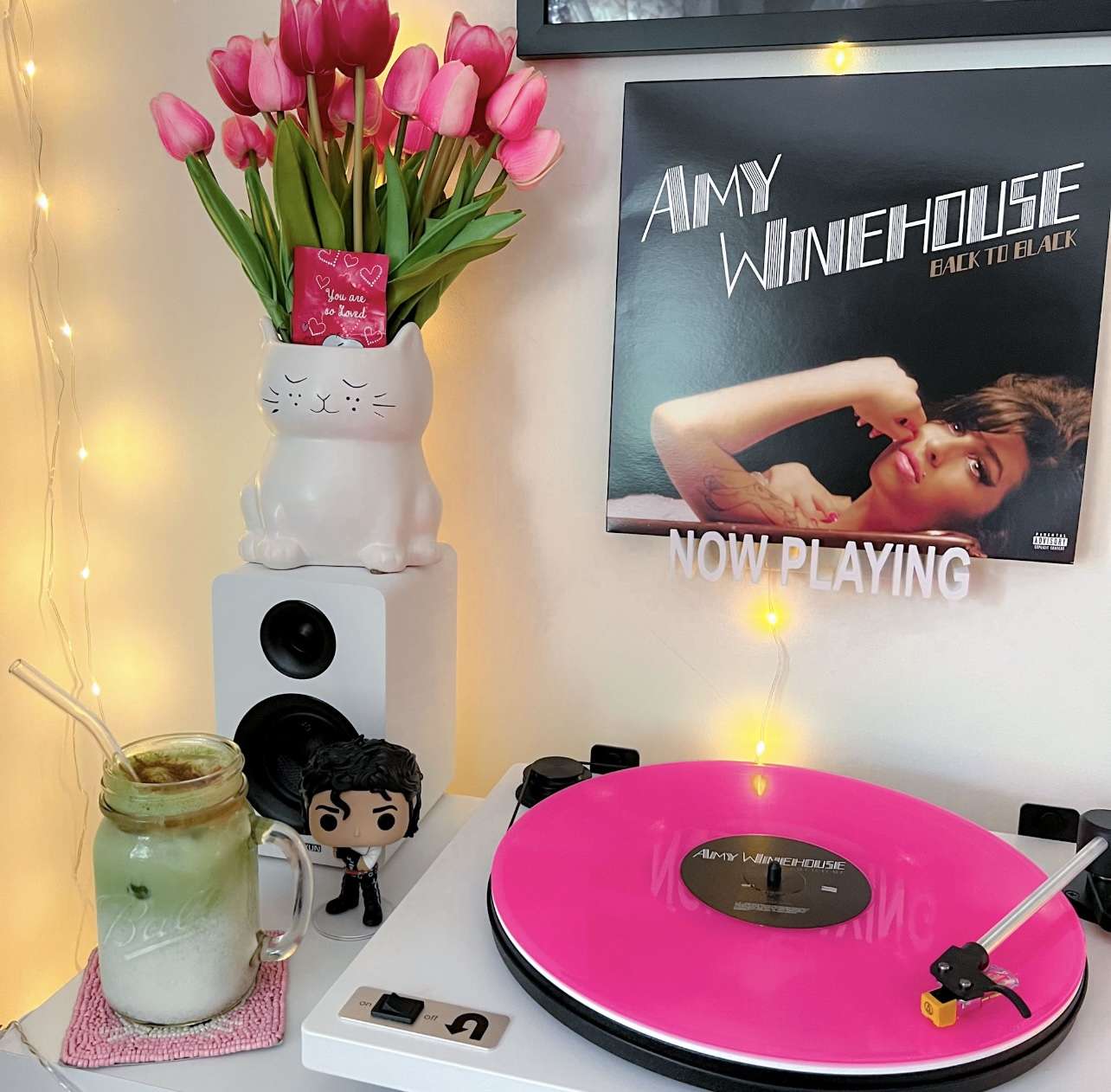 A cozy vinyl setup with a white turntable playing a bright pink record. Next to it is a small Funko Pop figurine, a cat-shaped vase with pink tulips, and white bookshelf speakers. On the wall is Amy Winehouse’s Back to Black album cover with a “Now Playing” sign below it, accented by string lights.