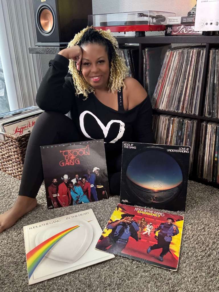 A woman sits on the floor in front of her vinyl shelves, surrounded by Kool & the Gang records. She leans against the record cabinet smiling, with four albums spread out on the carpet, including Something Special and Emergency. A turntable and speaker are visible behind her.