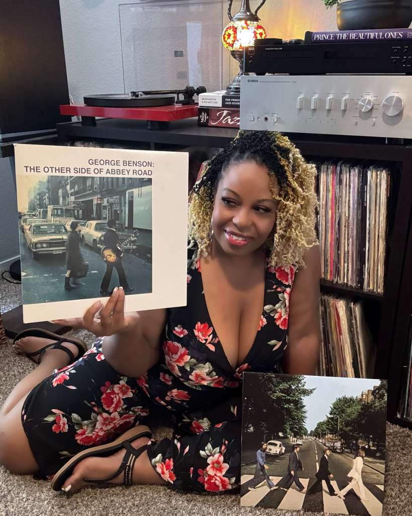 A woman in a floral dress sits on the floor next to her record collection, holding George Benson’s The Other Side of Abbey Road album. A copy of The Beatles’ Abbey Road album sits on the carpet in front of her. A turntable and stereo system are visible in the background.