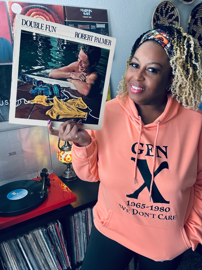 A woman wearing a peach hoodie that reads “GEN X 1965–1980 We Don’t Care” stands by her vinyl setup, smiling while holding Robert Palmer’s Double Fun album. A record spins on the turntable in the background.