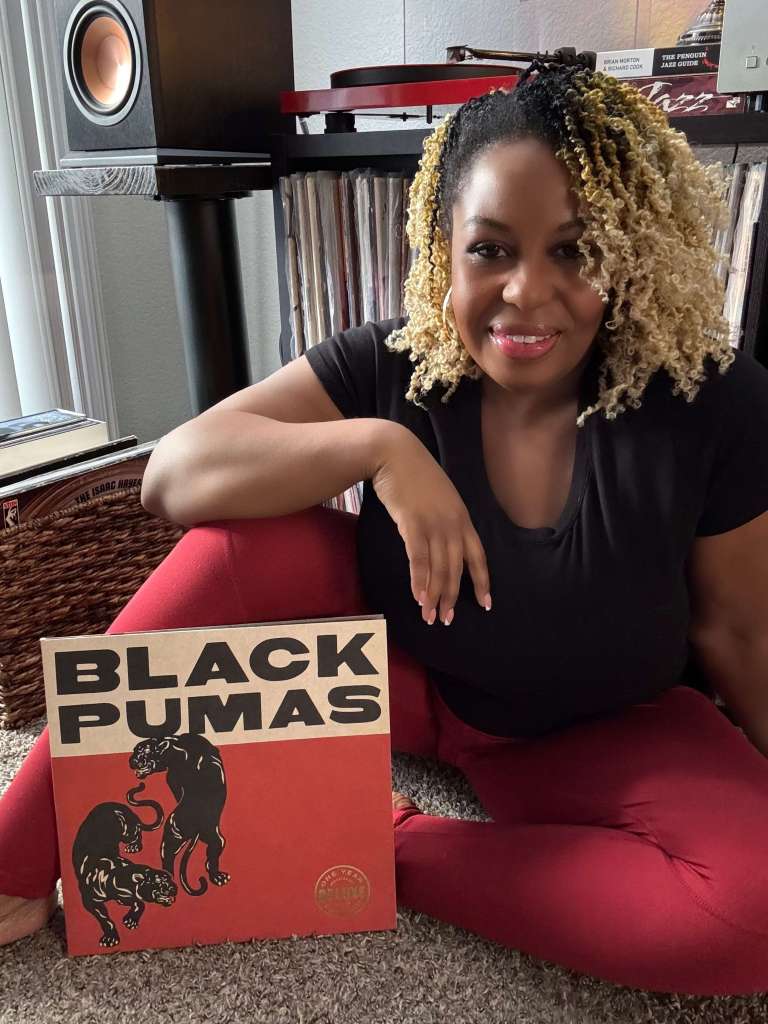 A woman sits cross-legged on the floor beside her record collection, holding a Black Pumas vinyl record. She is smiling, wearing a black top and red pants, with a turntable and bookshelf speakers in the background.