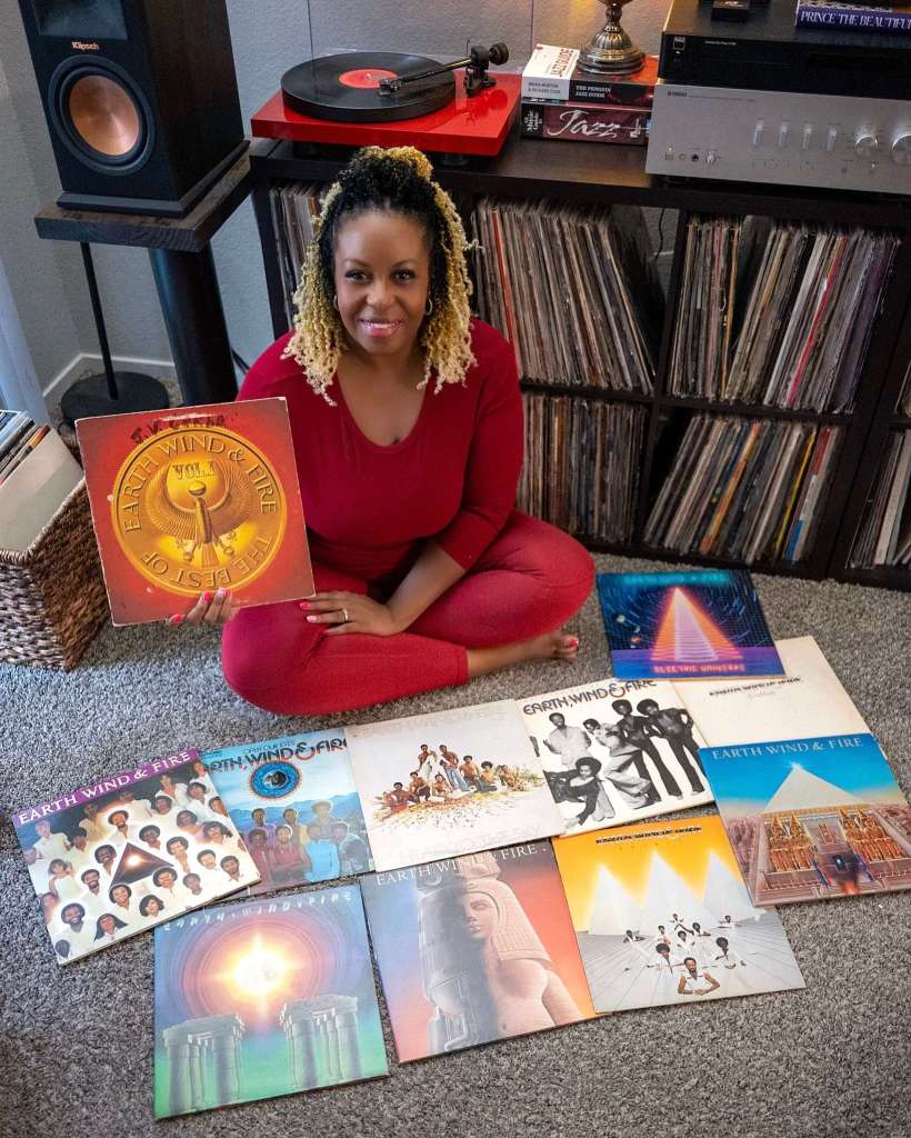 A woman in red sits cross-legged on the carpet, holding Earth, Wind & Fire’s The Best of Vol. 1 album. Several more Earth, Wind & Fire records are spread across the floor in front of her, with her record collection and setup in the background.