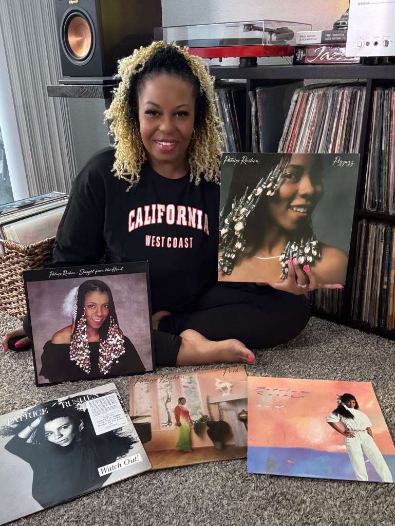 A woman sits in front of her vinyl collection, surrounded by Patrice Rushen records. She holds one album cover up while several others are spread neatly across the carpet. A red turntable and stereo setup are visible behind her.