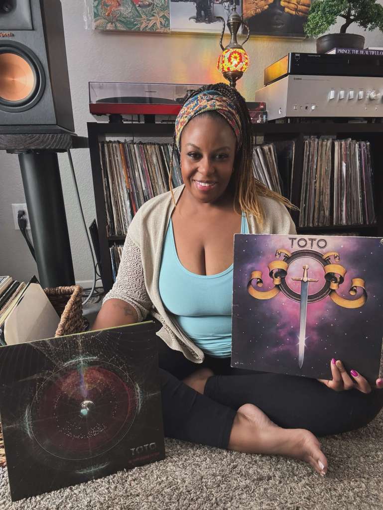 A woman sits cross-legged on the floor in front of her record collection, holding a Toto album with a sword on the cover. Another Toto record leans against her leg. She wears a light cardigan over a turquoise top, with speakers, a turntable, and stereo gear visible in the background.