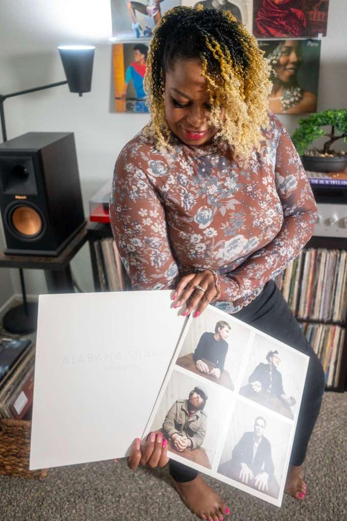 A woman smiles while looking down at an open Alabama Shakes Boys & Girls vinyl. She is barefoot, with bright pink nails, standing near her record collection and turntable setup, with album covers displayed on the wall.