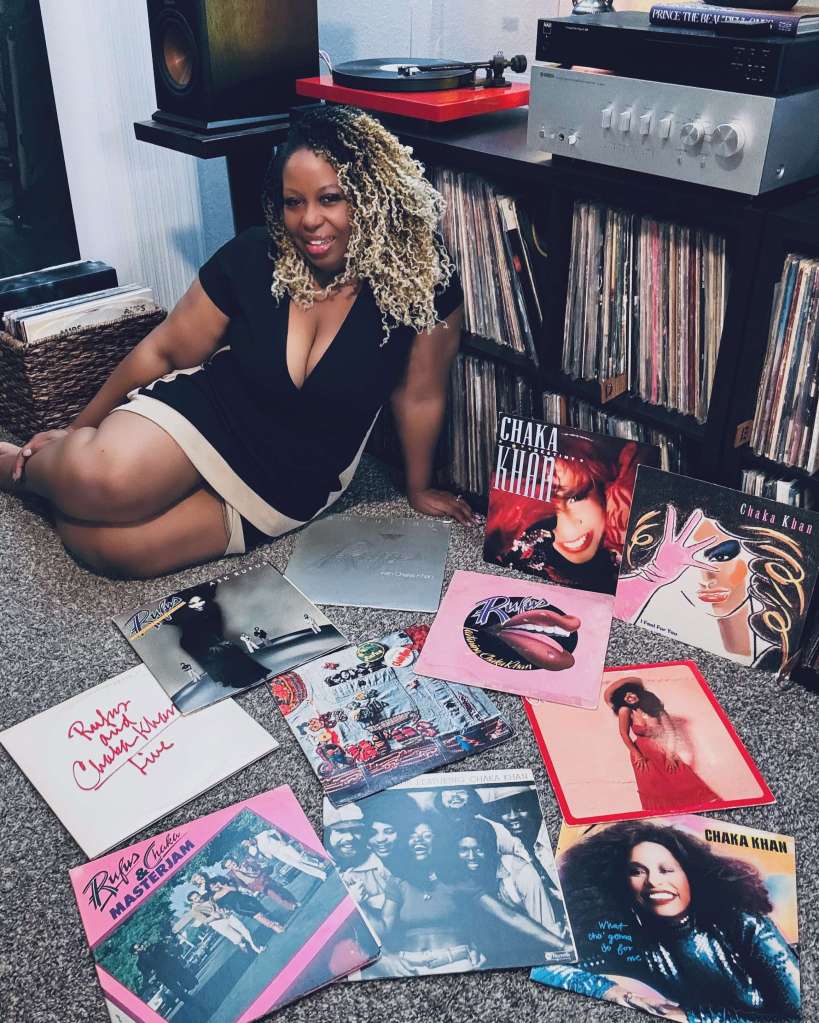 A woman sits on the floor in front of her stereo setup, smiling with a collection of Chaka Khan and Rufus vinyl records spread across the carpet. She wears a black dress with beige trim, and a turntable spins in the background.