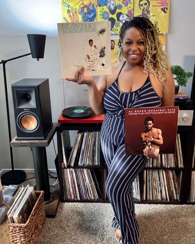 A woman stands beside her vinyl collection, smiling and holding two Al Green records: I’m Still in Love with You and Greatest Hits. A turntable, stereo, and bookshelf speakers sit behind her, with colorful album covers on the wall.