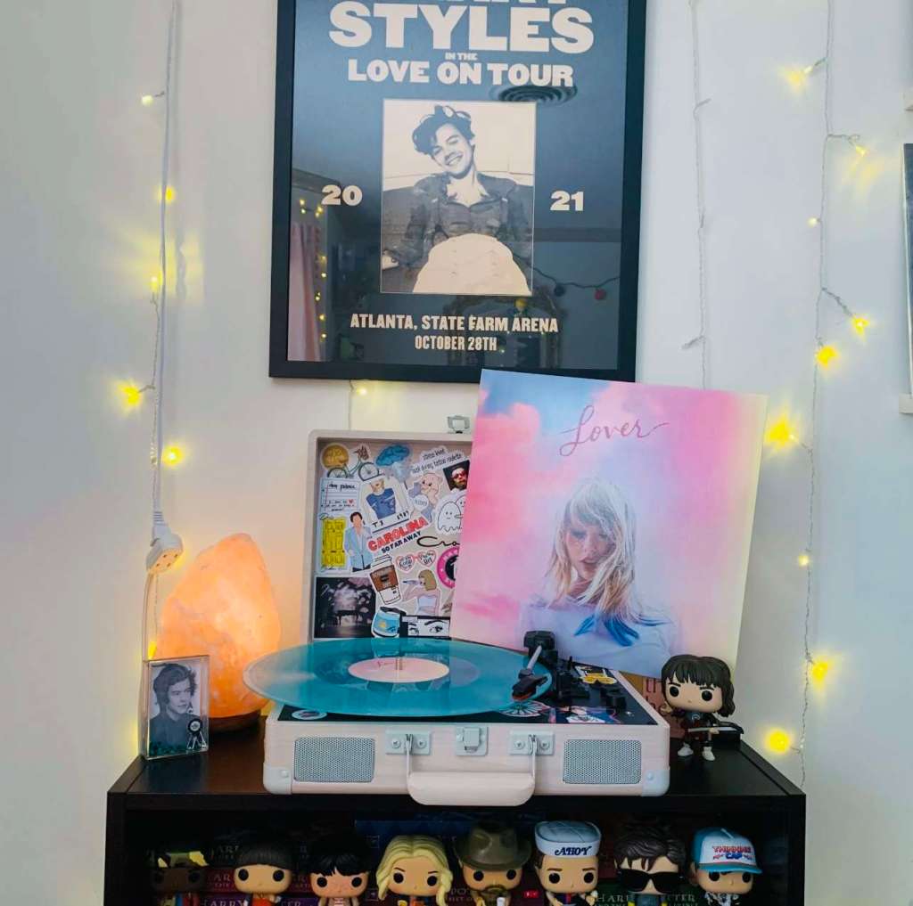 Turntable setup with a translucent blue Taylor Swift “Lover” vinyl on the platter, displayed beneath a Harry Styles tour poster and surrounded by Funko Pop figures and string lights.