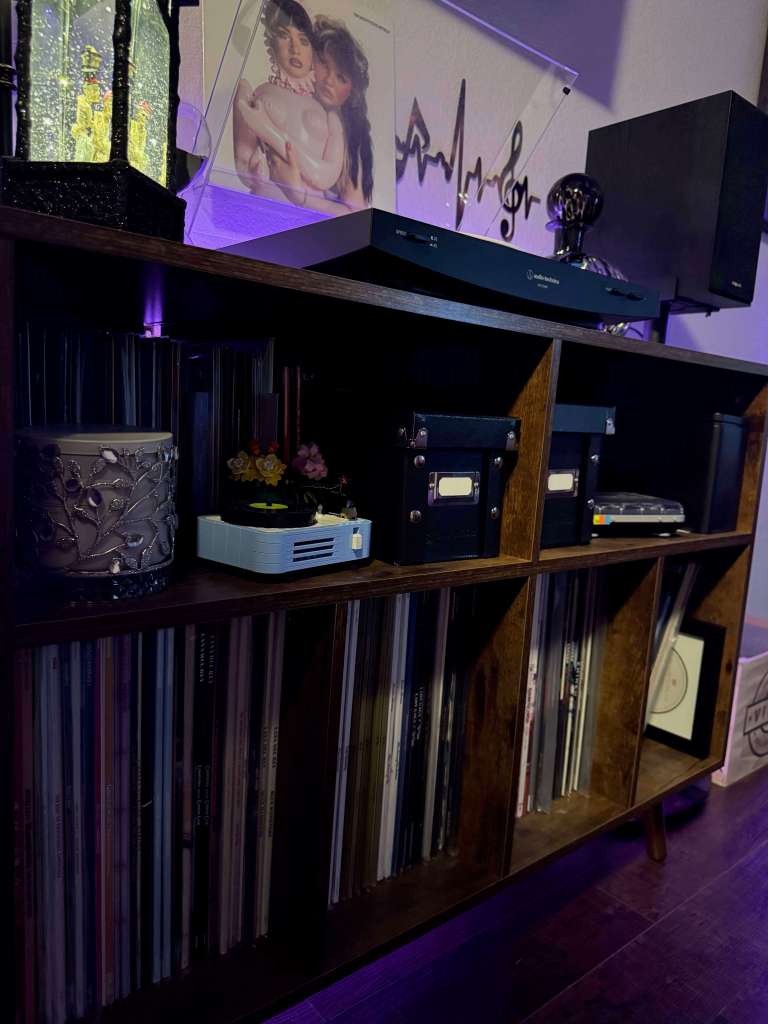 Dark wood vinyl storage shelf with six compartments filled with records, small decor pieces on top including a jeweled candle holder, LEGO flower model, and black storage boxes. Above the shelf sits an Audio-Technica turntable and black bookshelf speaker, lit by purple ambient lighting with an album cover displayed in a clear stand.