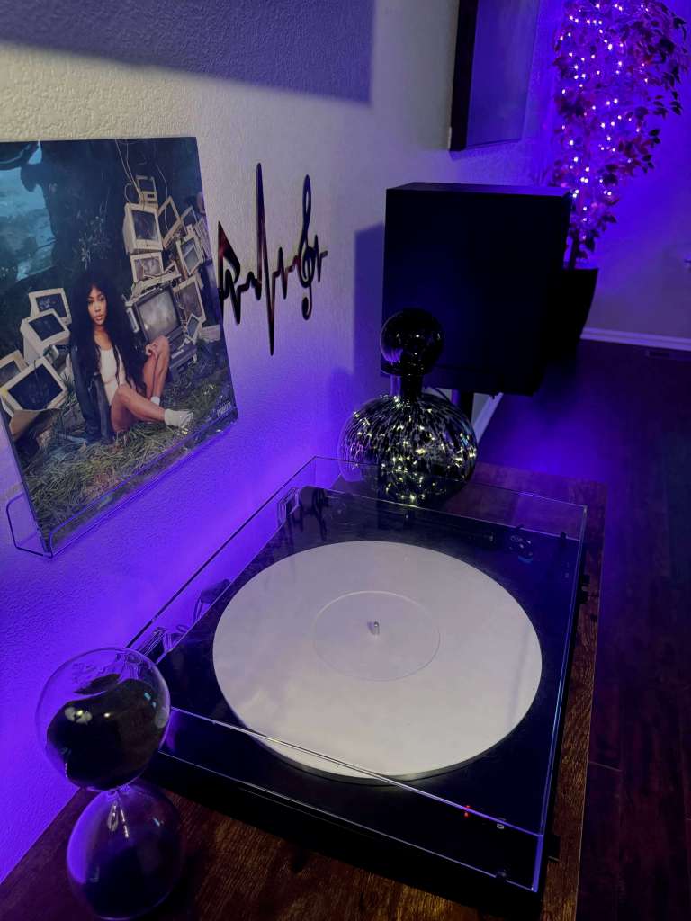 Vinyl listening setup under purple LED lights, featuring a white vinyl record spinning on a clear-platter turntable. A framed SZA album cover hangs on the wall with a treble clef heartbeat decal beside it. A decorative glass orb and black hourglass sit on the wooden cabinet, with a black speaker in the background.
