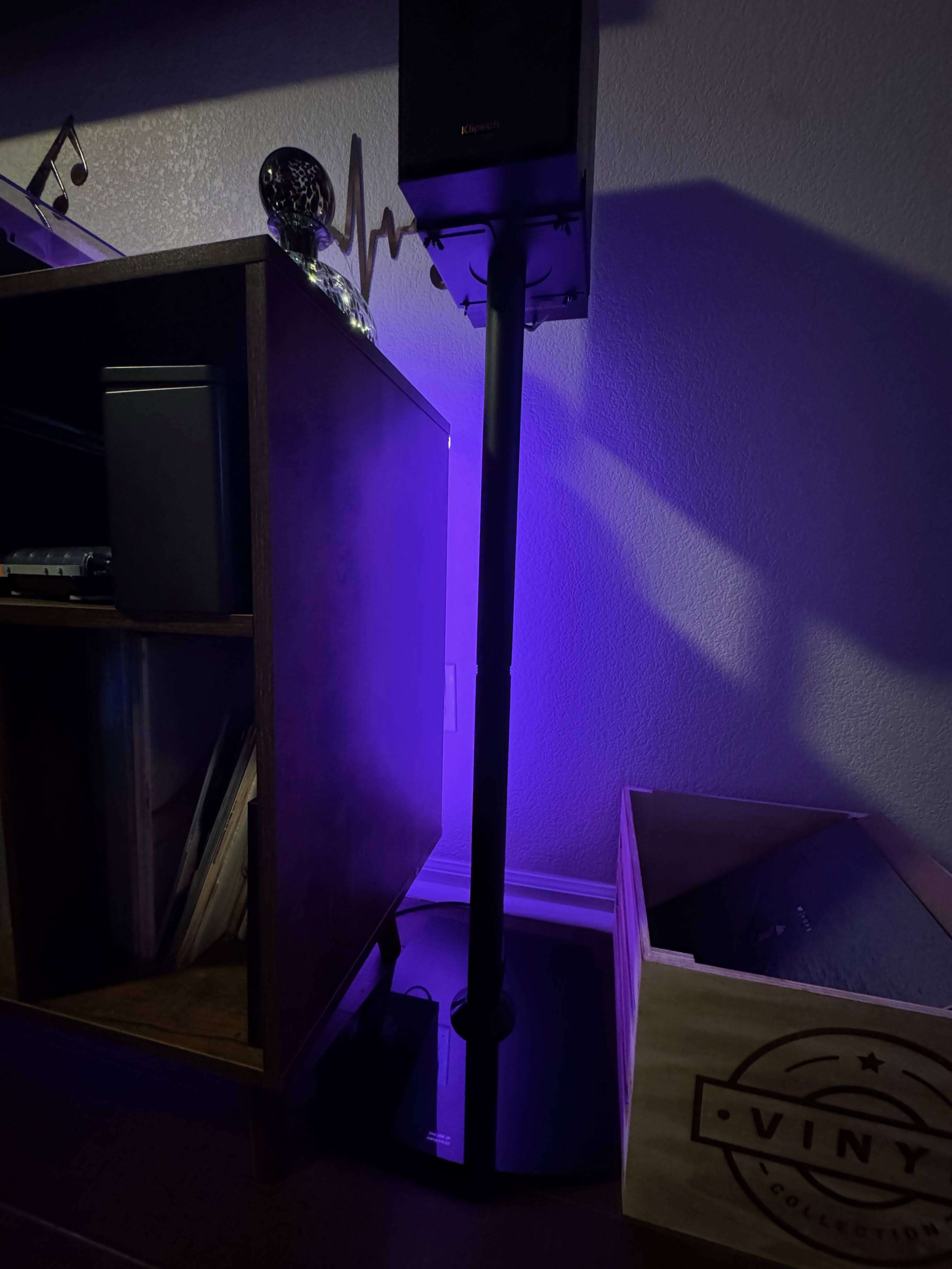 Close-up of a black Klipsch bookshelf speaker mounted on a tall stand beside a dark wood vinyl storage cabinet. The setup is accented by purple LED backlighting, with a glass decor piece on top of the cabinet and a wooden crate labeled “Vinyl Collection” on the floor holding records.