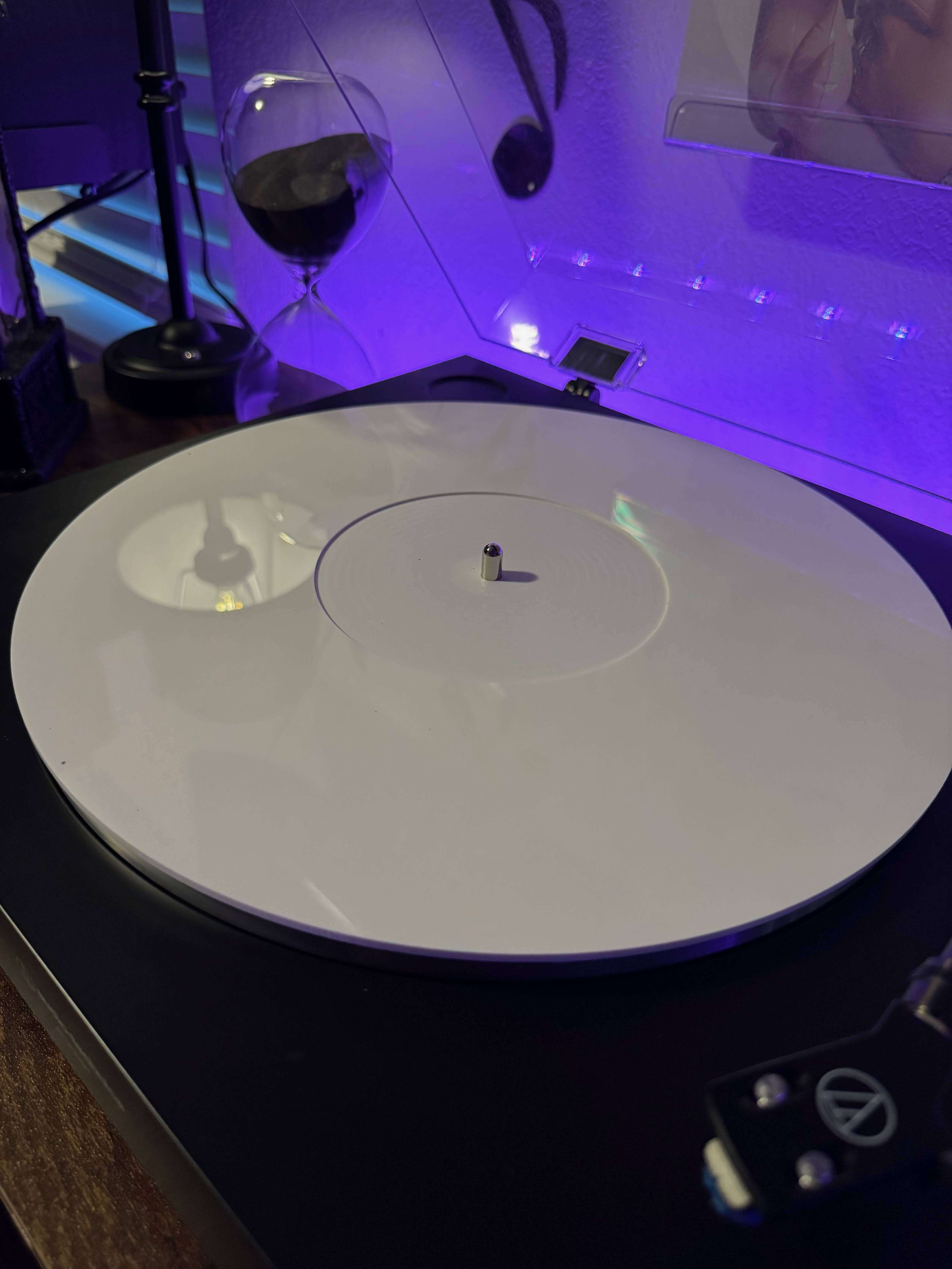Minimalist white vinyl record on a black turntable with a clear dust cover, paired with a black sand hourglass and glowing purple accent lighting.