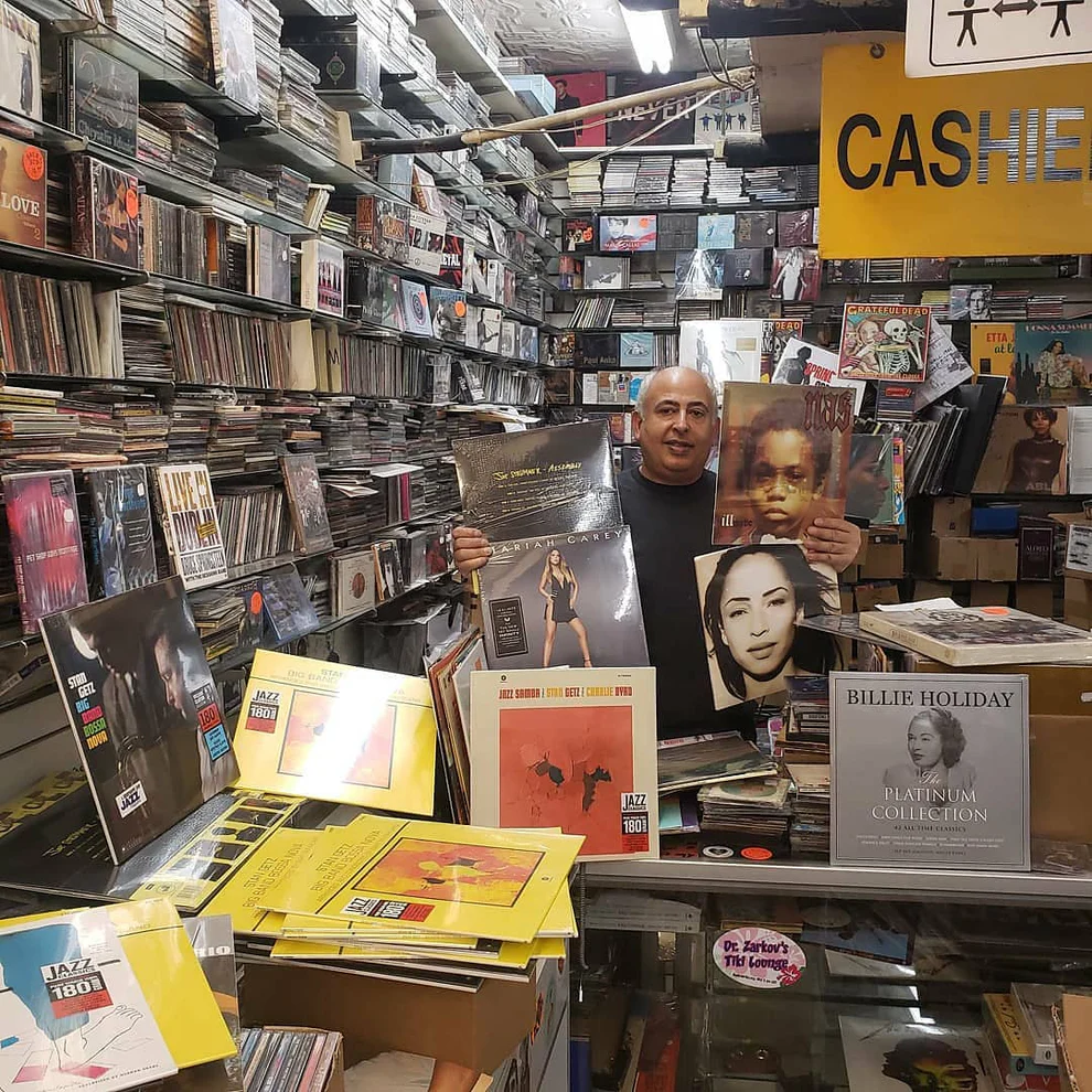 A record shop owner stands behind a crowded counter, holding up several vinyl records including albums by Mariah Carey, Sade, and Lauryn Hill. The store is packed floor-to-ceiling with CDs, vinyl, and posters, creating a dense, eclectic atmosphere. A yellow “Cashier” sign hangs above, and a Billie Holiday record is prominently displayed in front.