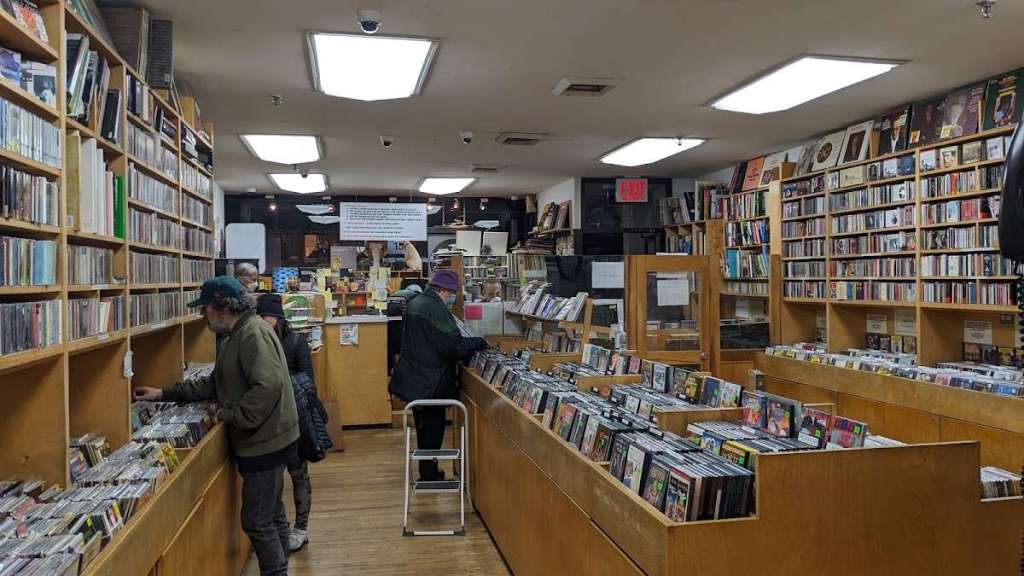 Interior of Academy Records & CDs, with tall wooden shelves stacked with CDs and vinyl. Several customers browse the aisles, while fluorescent ceiling lights brighten the space. Rows of records and CDs fill the shop, creating a dense, organized atmosphere.