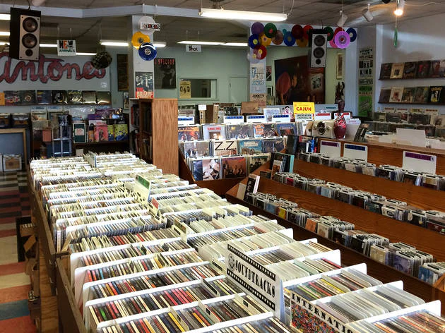 Interior of Antone’s Record Shop, with rows of bins filled with vinyl records organized by genre. Signs mark sections like “Soundtracks” and “Cajun,” while album covers and posters decorate the walls. The colorful tiled floor and ceiling decorations made of vinyl records add to the vintage, eclectic atmosphere.