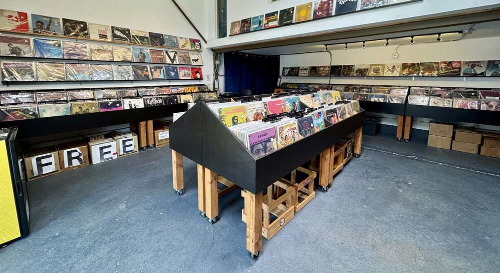 Interior of a minimalist record store with black bins of vinyl records arranged on wooden stands. Album covers line the walls in neat rows, and cardboard boxes labeled “FREE” sit underneath the shelves. The concrete floor and simple layout give the space an industrial, no-frills feel.