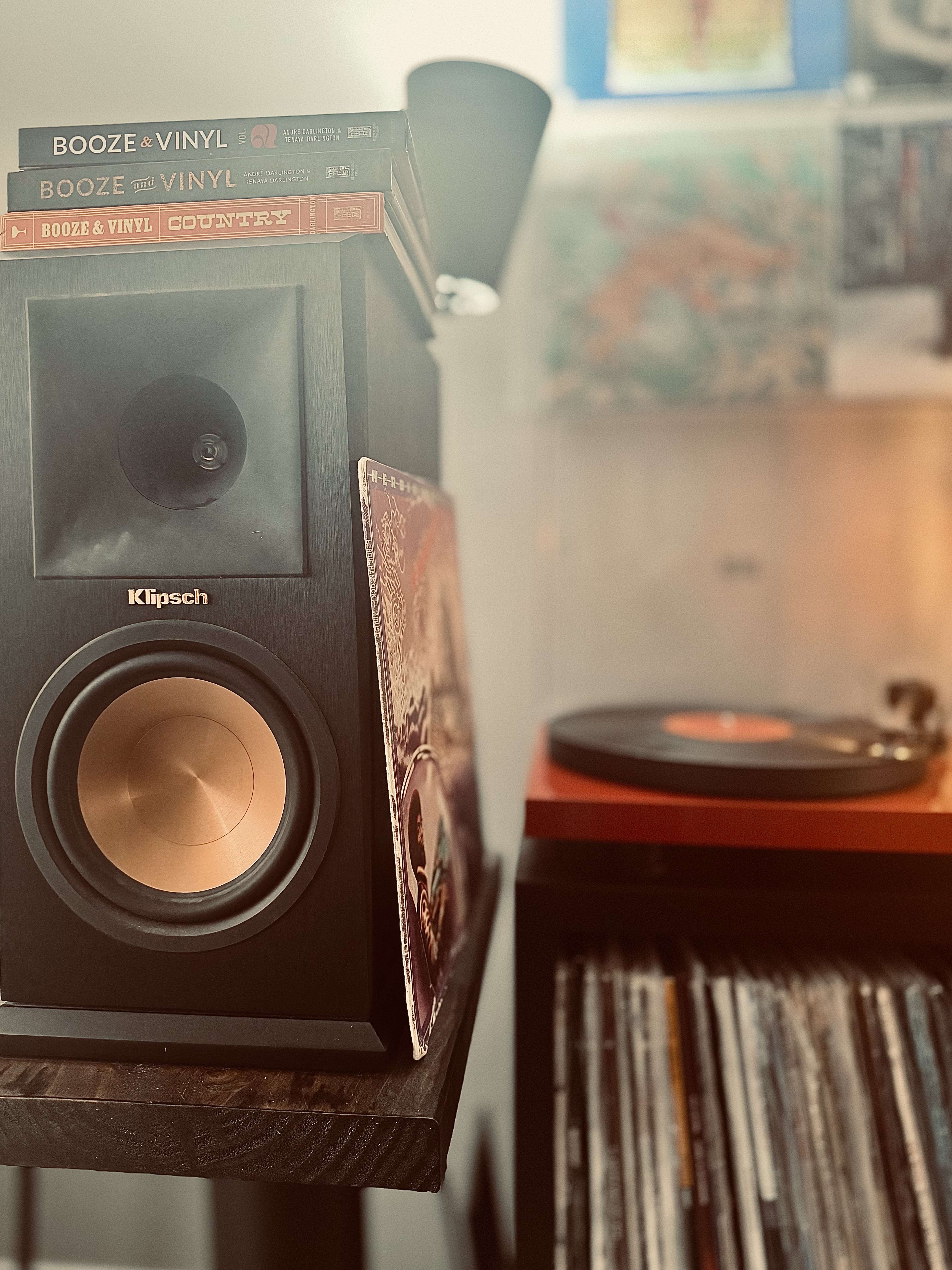 A black Klipsch bookshelf speaker with a copper cone, topped with three Booze & Vinyl books. A vinyl record spins on a red turntable in the background, with a record shelf below.