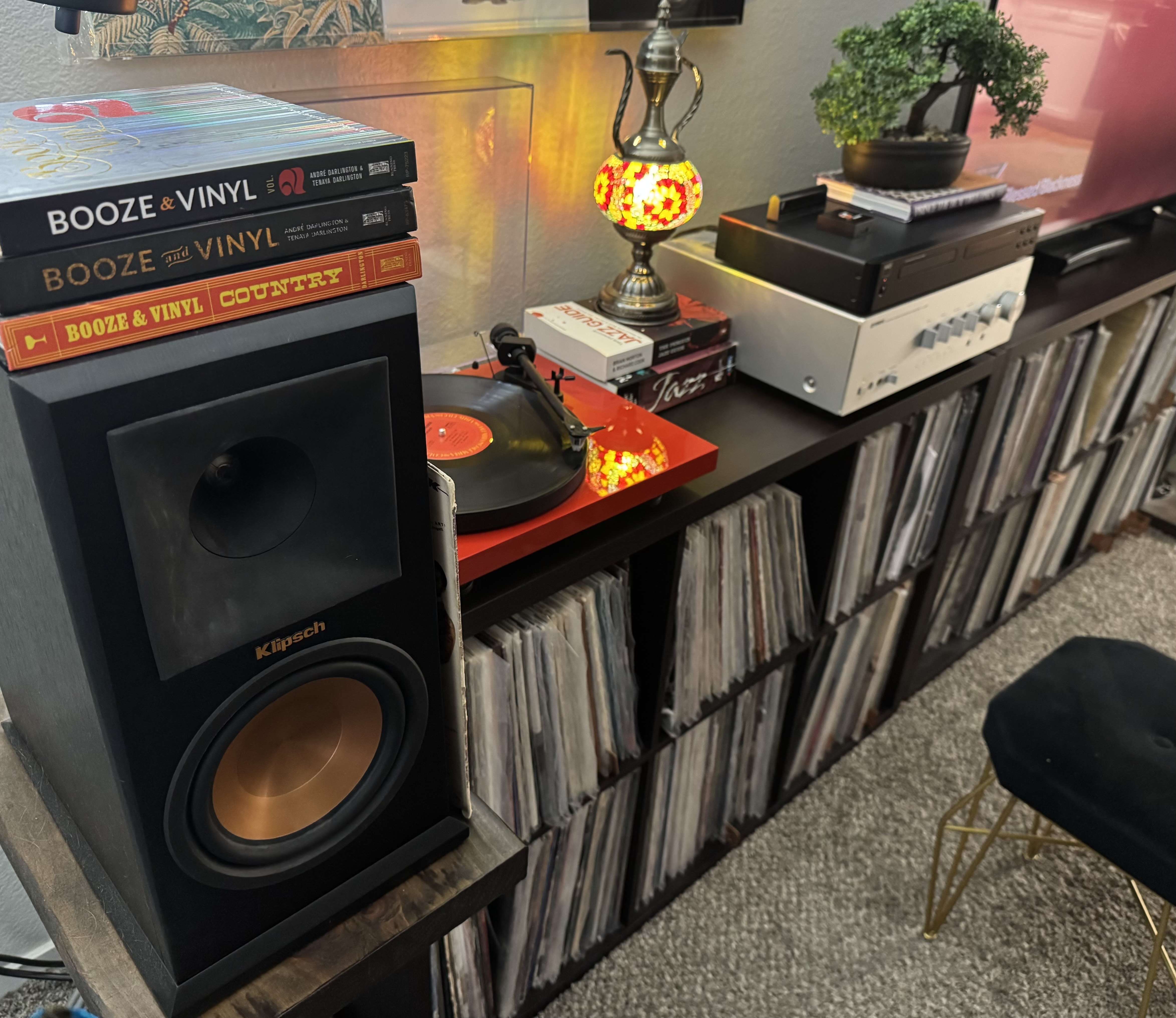 A black Klipsch bookshelf speaker with a copper cone, topped with three Booze & Vinyl books. A vinyl record spins on a red turntable in the background, with a record shelf below.