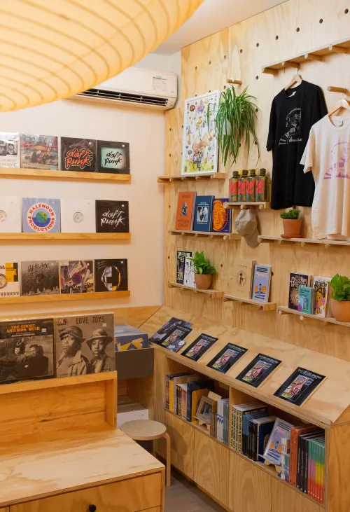 A modern record store corner with light wood shelving, vinyl records displayed on the wall, books and zines on angled racks, and potted plants adding greenery. Two graphic t-shirts hang on the wall above, alongside colorful posters and merch, creating a cozy and curated atmosphere.